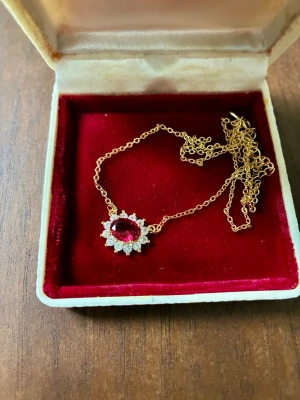 Halsband i silver med röd sten - Ett vackert halsband med en gnistrande röd sten (vet ej vad för sten) omgiven av små kristaller. Halsbandet är tillverkat av silver, stämplat S925. Levereras i ask från Rappl, Uhren-Juwelen, Wiesbaden.  Själva hänget är ca 15 x 13 mm stort och kedjan är 40 cm lång med möjlighet att förlänga några centimeter.  Jag vet dessvärre inte ursprung, värde eller övriga detaljer på smycket, men har aldrig haft anledning att tro att det är alltför dyrt eller exklusivt. Säljes för att det inte är min stil o