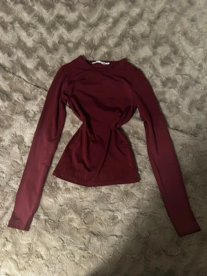 Mörkröd långärmad topp - Snygg mörkröd långärmad topp med figurnära passform och rund halsringning. Tillverkad i ett stretchigt material som sitter tight och markerar midjan. Perfekt att styla med jeans eller kjol för en trendig look.