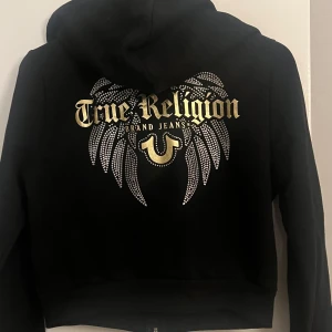 Svart kroppad hoodie True Religion med bling - Svart kroppad hoodie från True Religion med dragkedja och huva. På ryggen finns stora vingar i silver och guld med bling och logga i guldtext. Liten logga framtill i guld. Snygg streetwear-vibe och skön passform. Perfekt för dig som gillar detaljer och vill sticka ut.