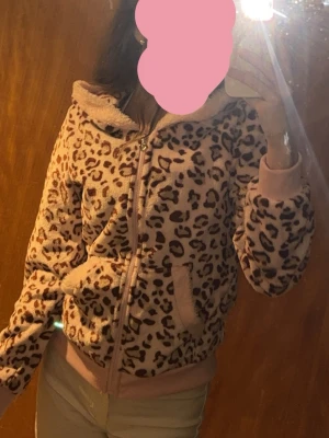 Rosa Leopardmönstrad fluffig hoodie - Casual damtröja med leopardmönster och dragkedja med ett hjärta. Väldigt skönt och mjukt material, luktar lite plast men tvättas då den är ny. Säljer för att den är för kort i ärmarna. Är man lika lång som mig (1,70cm) så skulle M pa
