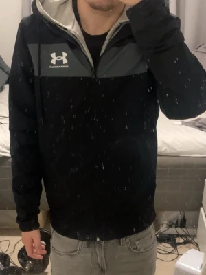 Svart vindjacka från Under Armour - Svart vindjacka med grått parti över bröstet och vit Under Armour-logga. Jackan har huva med dragsko, hel dragkedja och elastiska muddar vid ärmsluten. Perfekt för blåsiga dagar och sportiga outfits.