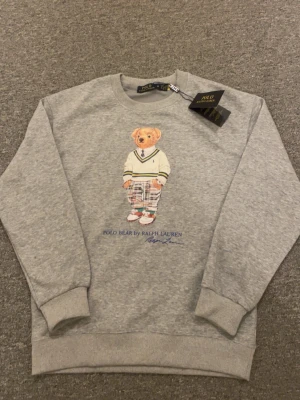 Grå sweatshirt Polo Bear Ralph Lauren - Grå sweatshirt från Ralph Lauren med ikoniska Polo Bear-trycket framtill. Tröjan har rund hals, ribbade muddar vid ärmslut och nederkant samt en avslappnad passform. Perfekt för en casual och trendig look.