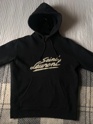 Svart Saint Laurent hoodie med tryck - Svart hoodie från Saint Laurent med vit logga i graffiti-stil på bröstet. Klassisk känguruficka framtill, dragsko i huvan och mjukt material. Perfekt för en avslappnad och trendig look.