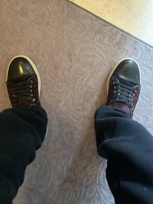 Lanvins glans - Snygga sneakers i vinrött mockaliknande material med svart tåparti i läderlook och vita sulor. Skorna har svarta skosnören och metallfärgade öljetter. Klassisk låg modell som passar till många olika stilar.  Skorna är använda men inte trasiga ( trasigt inuti skon vid hälen, slitna skosnören) 