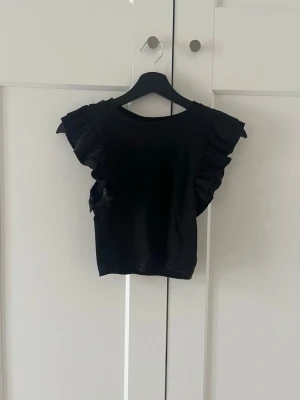 Svart topp med volangärmar - Snygg svart topp med volangdetaljer på ärmarna. Toppen har rund halsringning och är ärmlös med breda volanger som ger en cool och trendig look. Perfekt att styla med jeans eller kjol för en modern outfit.Säljer pga för liten.