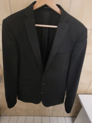 Kavaj från Tiger of Sweden - Säljer en mycket stilren och välskuren kavaj från Tiger of Sweden, tillverkad i högkvalitativt ulltyg från det anrika italienska väveriet Angelico. Kavajen är framtagen i ett exklusivt samarbete ("Exclusively for Tiger of Sweden") vilket innebär en fantastisk lyster, hög andningsförmåga och ett tyg som håller formen extremt väl. Detaljer: Märke: Tiger of Sweden Tyg: 100 % Ull (Lanificio Angelico, Italy)