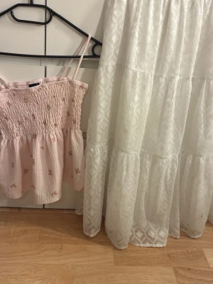 Ljusrosa smockad linnetopp med blommor - Supersöt ljusrosa linnetopp med smockad överdel och tunna axelband. Toppen har små broderade blommor och volang nedtill, perfekt för en somrig look. Lätt och luftig känsla.