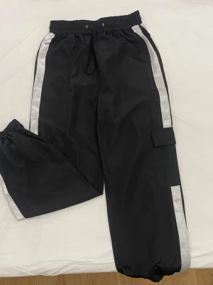 Svarta cargobyxor med vita ränder - Säljer ett par svarta cargobyxor med breda vita ränder längs sidorna. Byxorna har resår i midjan med dragsko, ficka på benet och resår vid bensluten. Perfekta för en avslappnad och sportig stil.