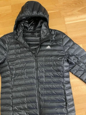 Svart dunjacka med huva från Adidas - Snygg svart dunjacka från Adidas med huva och dragkedja framtill. Jackan har ett quiltat mönster och Adidas-logga på bröstet. Perfekt för kalla vinterdagar och enkel att matcha med olika outfits.