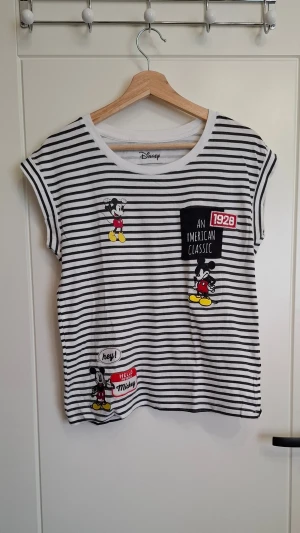 Randig Mickey Mouse t-shirt Disney - Säljer en vit och svart randig t-shirt från Disney med flera tryck av Mickey Mouse och textdetaljer. T-shirten har korta ärmar, rund halsringning och är croppad i modellen. Märkt med Cropp och har coola broderade detaljer framtill.