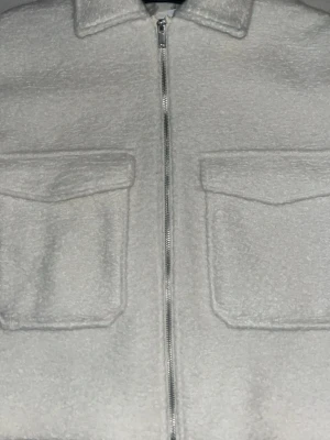 Vit teddyjacka med fickor och dragkedja - Mysig vit teddyjacka med två stora bröstfickor med lock och dragkedja framtill. Jackan har klassisk krage och är tillverkad i fluffigt teddy-material som ger en cool och avslappnad look.