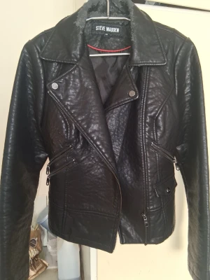 Svart bikerjacka från Steve Madden - Snygg svart bikerinspirerad skinnjacka från Steve Madden med klassisk krage, sned dragkedja och flera dragkedjefickor. Jackan har en cool, glansig finish och detaljer som ger en edgy look. Perfekt för dig som vill sticka ut med stil.