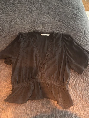 Svart omlottblus från Zara - Snygg svart omlottblus från Zara med spetsdetaljer längs v-ringningen och midjan. Blusen har korta ärmar och ett lätt transparent tyg med diskret prickmönster. Perfekt att styla med jeans eller kjol för en trendig look.