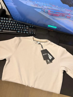 Beige sweatshirt från BOSS - Stilren beige sweatshirt från BOSS med rund halsringning och diskreta ribbade muddar. Tröjan har en enkel design med en liten randig detalj på insidan av kragen. Perfekt för en avslappnad och trendig look. Pris kan diskuteras!!