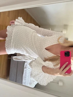 Vit kort klänning med spetsvolang - Superfin vit kort klänning från AG x NA-KD med v-ringning och långa ärmar. Klänningen har spetsvolanger vid ärmslut och nederkant samt ett broderat hålmönster över hela plagget. Perfekt för sommarens alla tillfällen!