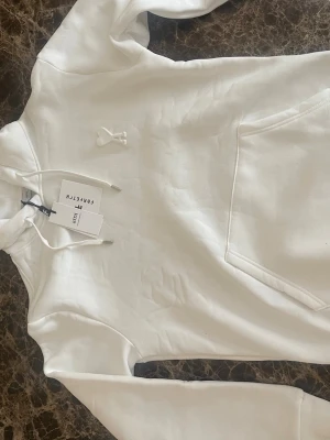 Vit hoodie  - Säljer en stilren vit hoodie från … med deras ikoniska hjärta-A broderat på bröstet. Hoodien har huva med dragsko och en stor magficka. Tillverkad i mjuk bomull för maximal komfort och en clean look.