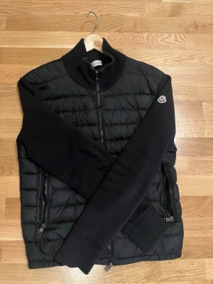 Svart pufferjacka från Moncler - Snygg svart pufferjacka från Moncler med quiltad front och stickade ärmar. Jackan har hög krage, två dragkedjefickor och Moncler-logga på ärmen. Perfekt för kalla dagar och riktigt trendig look.