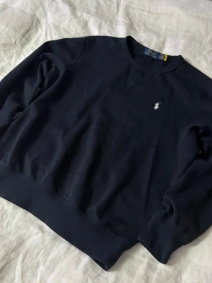 Mörkblå sweatshirt från Polo Ralph Lauren - Klassisk mörkblå sweatshirt från Polo Ralph Lauren med rund halsringning och broderad logga på bröstet. Tröjan har en relaxed fit och är tillverkad i mjuk bomull, perfekt för en chill och stilren look.
