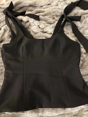 Svart linnetopp med knytband - Snygg svart linnetopp med breda axelband som knyts i rosetter på axlarna. Toppen har en rak passform och är perfekt att styla med jeans eller kjol. Enkel och stilren design som passar till många olika looks.