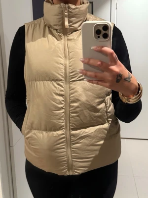 Beige dunväst med hög krage - Snygg beige dunväst med hög krage och dragkedja framtill. Västen har en quiltad design och två sidofickor. Perfekt att slänga över en hoodie eller tröja för extra värme. Enkel och stilren look sopassar till många outfits.