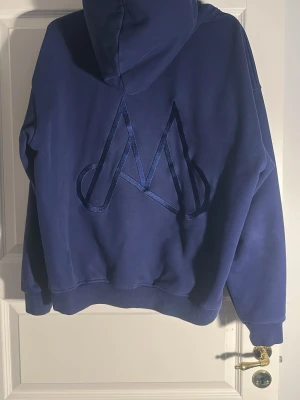 Blå hoodie med stor M-logga - Säljer en blå hoodie från Maggiore med stor M-logga i sammet på ryggen. Hoodien har huva, magficka och ribbade muddar vid ärmslut och nederkant. Tillverkad i mjukt bomullsmaterial som känns skönt mot huden.