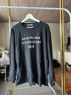 Svart långärmad topp Acne Studios - Svart långärmad topp från Acne Studios med tryck 'Acne Studios STOCKHOLM 1996' på bröstet. Klassisk rund halsringning och avslappnad passform. Perfekt för dig som gillar stilrena och trendiga plagg.