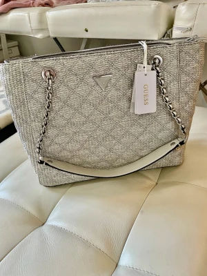 NY Guess Gandväska Giully Tote flera fack Silver - Snygg silver handväska från Guess med kedjedetaljer och quiltad struktur. Väskan har silverfärgade metalldetaljer, dragkedja upptill och ett Guess-emblem på framsidan. Materialet är textil med läderliknande inslag. Perfekt för att lyfta din outfit.  Storlek 45cm