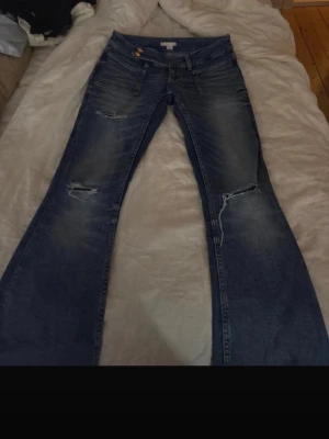 Blå bootcut jeans med slitningar - Säljer ett par blå bootcut jeans med slitningar på låren och knäna. Jeansen har klassisk femficksmodell, låg midja och snygg tvättad look. Perfekta för dig som gillar en avslappnad och trendig stil.