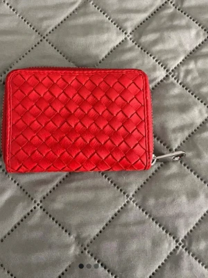 Röd plånbok - Snygg röd plånbok med flätad design och dragkedja runtom. Tillverkad i läder och har en kompakt, rektangulär form som gör den smidig att bära med sig. Perfekt för kort och kontanter.