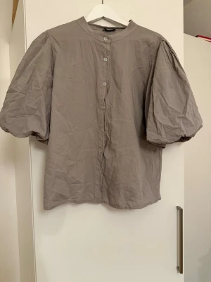 Grå blus med puffärm och knappar - Snygg grå blus med puffiga korta ärmar och knäppning framtill. Blusen har en rak passform och rund halsringning. Tillverkad i ett lätt och luftigt tyg som ger en avslappnad look.