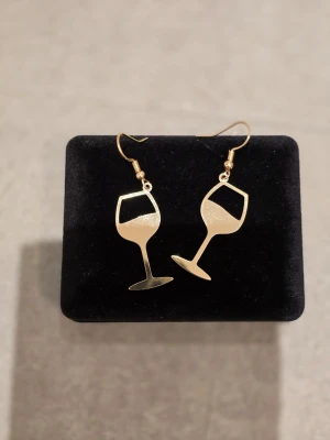 Guldiga örhängen med vinglasmotiv - Snygga örhängen i form av vinglas i guldfärgad metall. Perfekta för dig som vill sticka ut med en lekfull och trendig accessoar. Hängande modell med krokar som är enkla att bära. Passar dig som gillar unika smycken med humor.