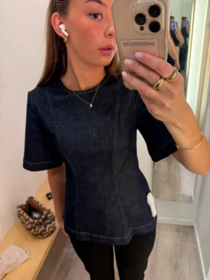 Mörkblå denimtop från Stockh LM Studio - Säljer en snygg mörkblå denimtop från Stockh LM Studio i storlek XS. Toppen har korta ärmar, markerade sömmar och rund halsringning. Materialet är denim och den har en diskret dragkedja i ryggen. Perfekt för dig som gillar en stilren och cool look.