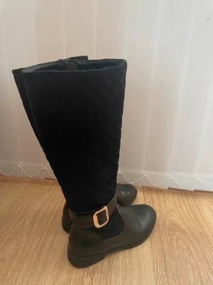 Svarta quiltade stövlar med spänne - Snygga svarta stövlar med quiltat skaft och dekorativt guldfärgat spänne vid ankeln. Stövlarna har rund tå och dragkedja på insidan för enkel påtagning. Materialet är en mix av textil och syntetläder som ger en cool och trendig look.