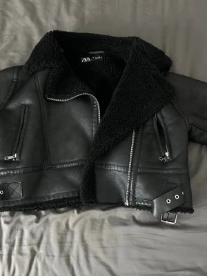 Svart skinnjacka från Zara - Cool svart skinnjacka från Zara med mjukt foder och bred krage. Jackan har flera silverfärgade dragkedjor, snedställda fickor och ett bälte längst fram. Använt fåtal gånger, säljs då den är för stor för mig.
