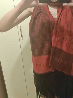 Röd Philipp Plein halsduk med döskalle - Snygg röd halsduk från Philipp Plein med all-over logomönster och stora döskallar i ändarna. Halsduken har fransar i kanterna och är tillverkad i ett mjukt material som känns lyxigt. Perfekt accessoar för att lyfta din outfit. Nypris 4500- 5000