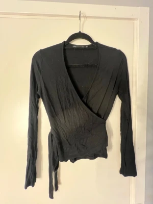 Svart omlottopp med knytband - Säljer en svart omlottopp med långa ärmar och knytband i midjan.  Storlek XS från lager 157 Perfekt att styla med jeans eller kjol för en trendig look. Skriv om du undrar något💕