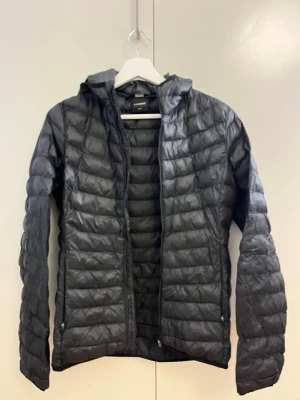 Svart pufferjacka från Everest - Snygg svart pufferjacka från Everest med huva och dragkedja framtill. Jackan har quiltad design och två sidofickor med dragkedja. Perfekt för kalla dagar och enkel att matcha med olika outfits. Tillverkad i lätt och värmande material.