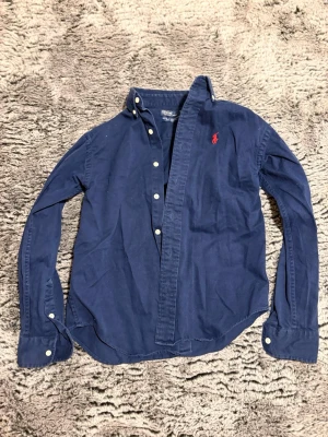 Marinblå skjorta från Polo Ralph Lauren passar XS - Passar XS Klassisk marinblå skjorta från Polo Ralph Lauren i 100% bomull. Skjortan har långa ärmar, knappar framtill och den ikoniska röda logotypen broderad på bröstet. Perfekt för en stilren och avslappnad look.