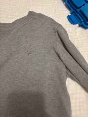 Grå stickad tröja från GANT - Snygg grå stickad tröja från GANT  med diskret logga broderad på bröstet. Tröjan har rund halsringning, långa ärmar och ribbade muddar vid  ärmslut och nederkant. Perfekt basplagg till garderoben. Bra skick hör av er vid mer info. Storlek 13-14 tests 158-164
