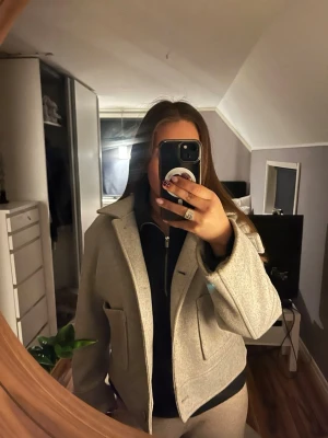 Ljusgrå ulljacka från 157 - Snygg ljusgrå jacka från 157 i ullblandning. Jackan har klassisk krage, stora fickor framtill och knäpps med knappar. Modellen är croppad och har en stilren, rak passform som passar perfekt till många outfits.