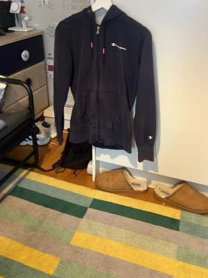 Mörkblå zip hoodie från Champion - Säljer en mörkblå hoodie från Champion med dragkedja framtill och två fickor. Klassisk Champion-logga broderad på bröstet och ärmen. Hoodien har huva med dragsko och ribbade muddar vid ärmslut och nederkant. Perfekt för en avslappnad streetwear-look.