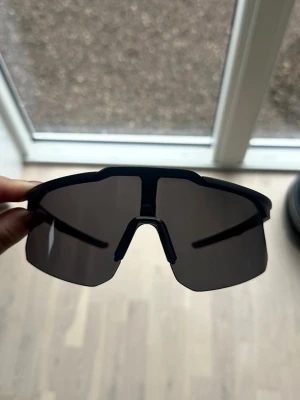 Svarta sportiga solglasögon - Snygga svarta solglasögon med breda, mörka linser och sportig design. Ramen är tunn och följer ansiktets form för en modern look. Perfekta för dig som vill ha en cool och futuristisk stil. Passar bra till löpning, i skidbacken eller liknande. UV-skydd finns. 