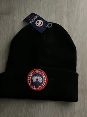 Svart mössa från Canada Goose - Stilren svart mössa från Canada Goose med klassisk uppvikt kant och det ikoniska runda märket framtill i rött, vitt och blått. Tillverkad i mjukt material som håller dig varm och passar perfekt till kalla dagar.