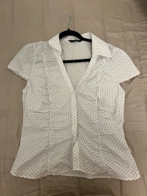 Vit prickig blus med krage - Snygg vit blus med svarta små prickar och klassisk krage. Storlek XL men passar M/L. Modellen har korta ärmar och knäppning framtill. Figurnära passform med lätt rynkade detaljer längs sidorna. Perfekt till jeans eller kjol för en trendig look.