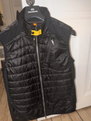 Svart dunväst från Parajumpers - Snygg svart dunväst från Parajumpers med quiltad design, hög krage och dragkedja framtill. Västen har en bröstficka med dragkedja och en gul detalj vid halsen. Perfekt för lager-på-lager och streetstyle. Äkta. Nypris runt 3800:- kvitto finns