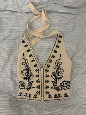 Beige broderad linnetopp med knyt - Trendig beige linnetopp med svart broderat blommönster och djup v-ringning. Toppen har knytband i nacken och smock baktill för skön passform. Perfekt för dig som vill ha en unik och snygg topp till sommaren. Storlek 40 MEN insydd till en s/m.