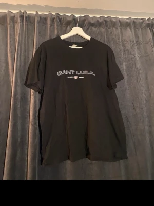 Svart GANT U.S.A. t-shirt - Snygg svart t-shirt från GANT med trycket 'GANT U.S.A.' och en liten sköld på bröstet. Klassisk passform och rund halsringning, perfekt till jeans eller shorts.