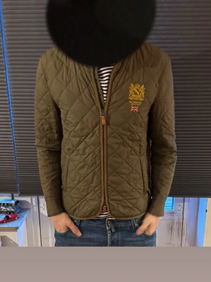 Olivgrön quiltad jacka från Morris - Snygg olivgrön quiltad jacka från Morris med bröstbrodyr och brittisk flagga. Jackan har dragkedja, två fickor fram och bruna detaljer vid kanterna. Perfekt för dig som vill ha en stilren och klassisk look.