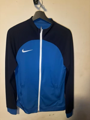 Blå och svart Nike träningsjacka - Nike träningsjacka i blått och svart med vit dragkedja och vit Nike-logga på bröstet. Jackan är i ett lätt och stretchigt material som passar perfekt till träning eller sport. Den är helt ny och har endast provat den 