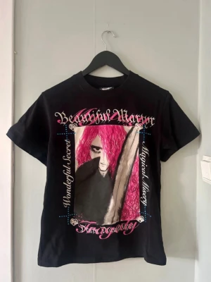 Bladee Martyr Tour Merch - Unik svart t-shirt med färgstarkt grafiskt tryck fram och bak i rosa, vitt och svart. Motivet har anime/vampyr-vibbar och cool text. T-shirten har normal passform och korta ärmar. Perfekt för dig som gillar edgy och konstnärlig stil.
